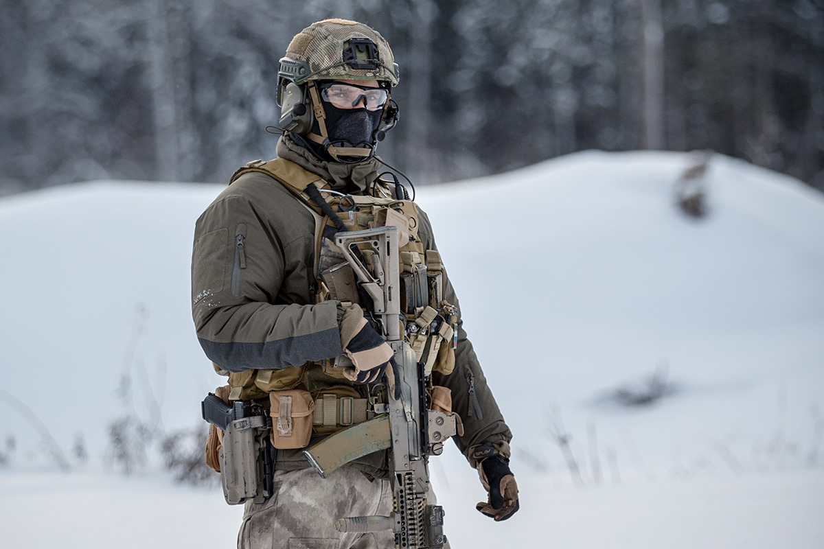 A security specialist in cold-weather protective equipment standing alert in an open snow-covered landscape.